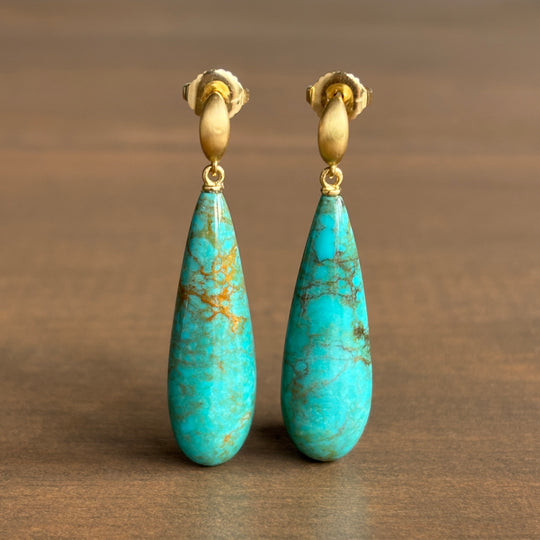 A pair of gold and turquoise drop earrings with teardrop-shaped turquoise stones and gold post backings, displayed on a brown wooden surface.