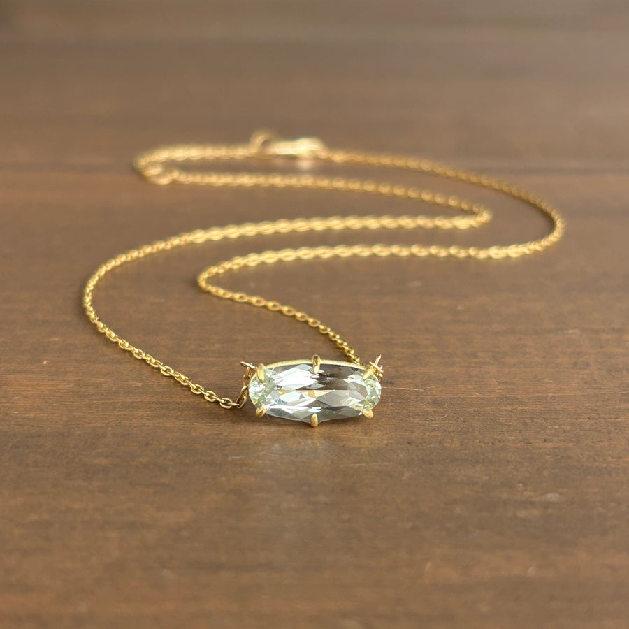 A gold chain necklace with a small oval clear gemstone pendant, displayed on a dark brown wooden surface.