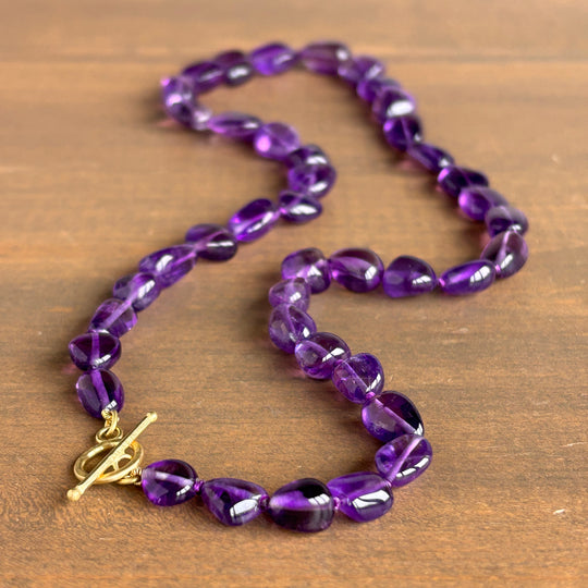 A beaded necklace made of glossy, faceted purple beads with a gold toggle clasp, displayed on a brown wooden surface.