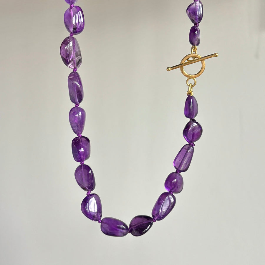 A beaded necklace made of polished purple amethyst gemstone beads with a gold toggle clasp, shown against a plain light gray background.