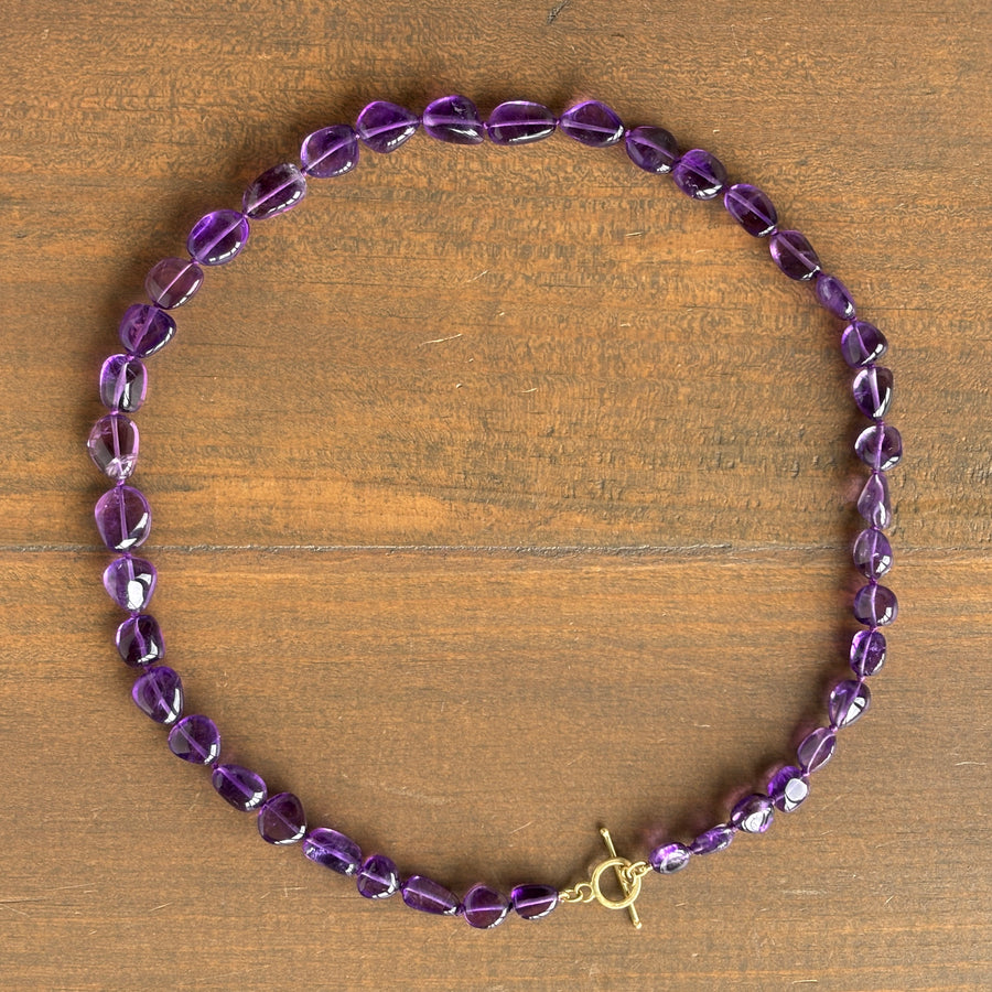 A beaded bracelet made of faceted purple amethyst beads with a gold toggle clasp, displayed on a brown wooden surface.