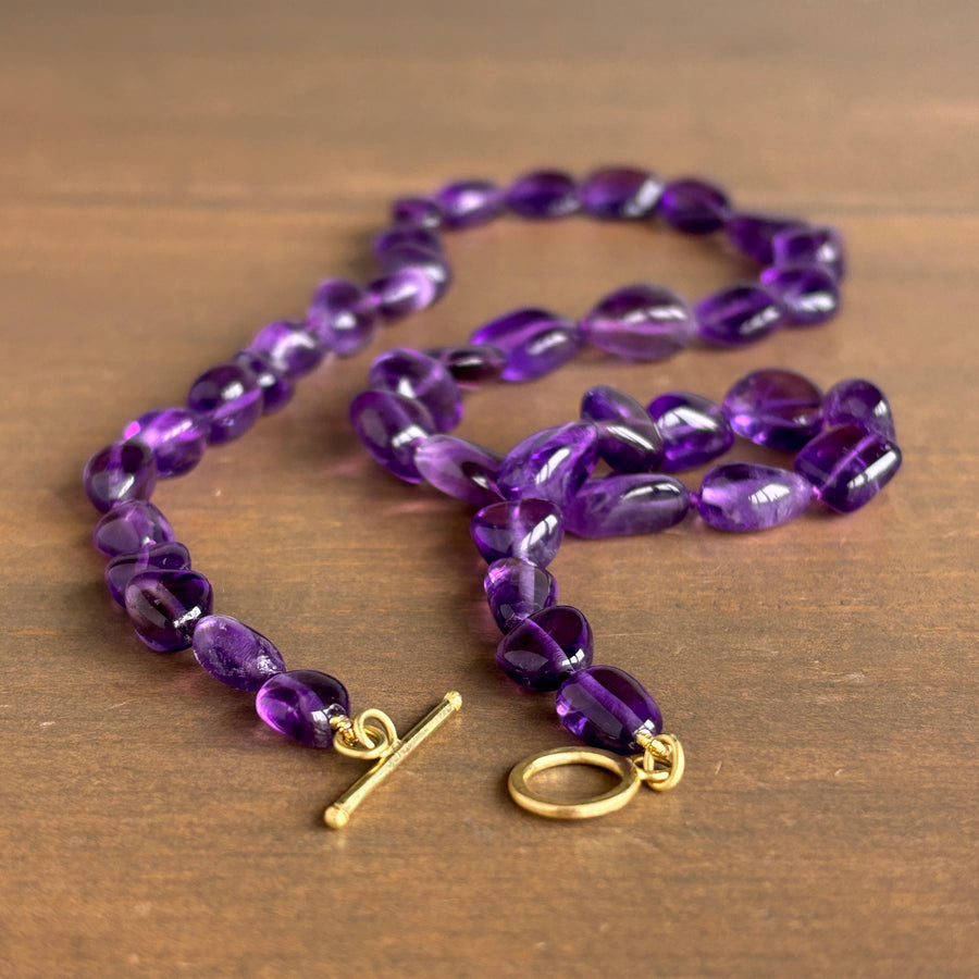 A beaded necklace made of polished purple amethyst teardrop beads with a gold toggle clasp, displayed on a brown wooden surface.