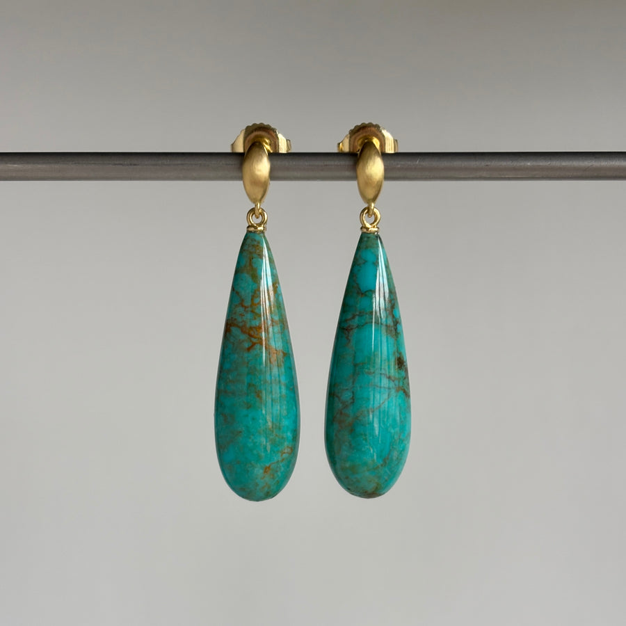 A pair of gold drop earrings with teardrop-shaped turquoise-blue stone pendants, hanging from a dark horizontal bar against a light gray background.