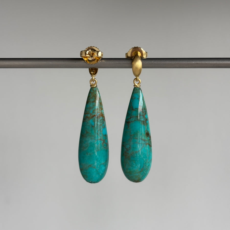 A pair of gold drop earrings with teardrop-shaped turquoise-blue stone pendants, hanging from a dark horizontal bar against a neutral gray background.