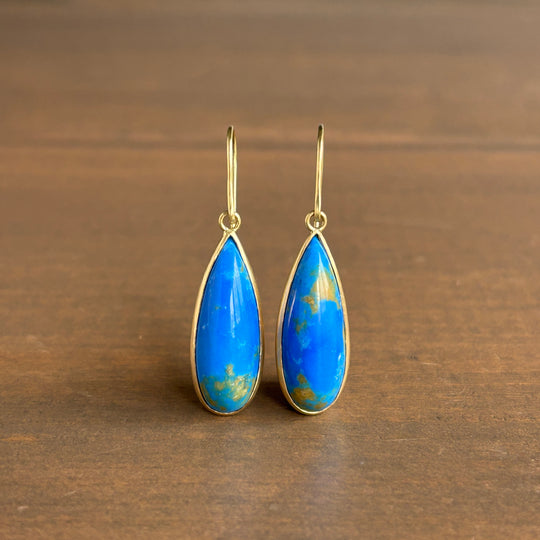 A pair of gold drop earrings with teardrop-shaped blue Ceruleite stones, displayed on a dark brown wooden surface.