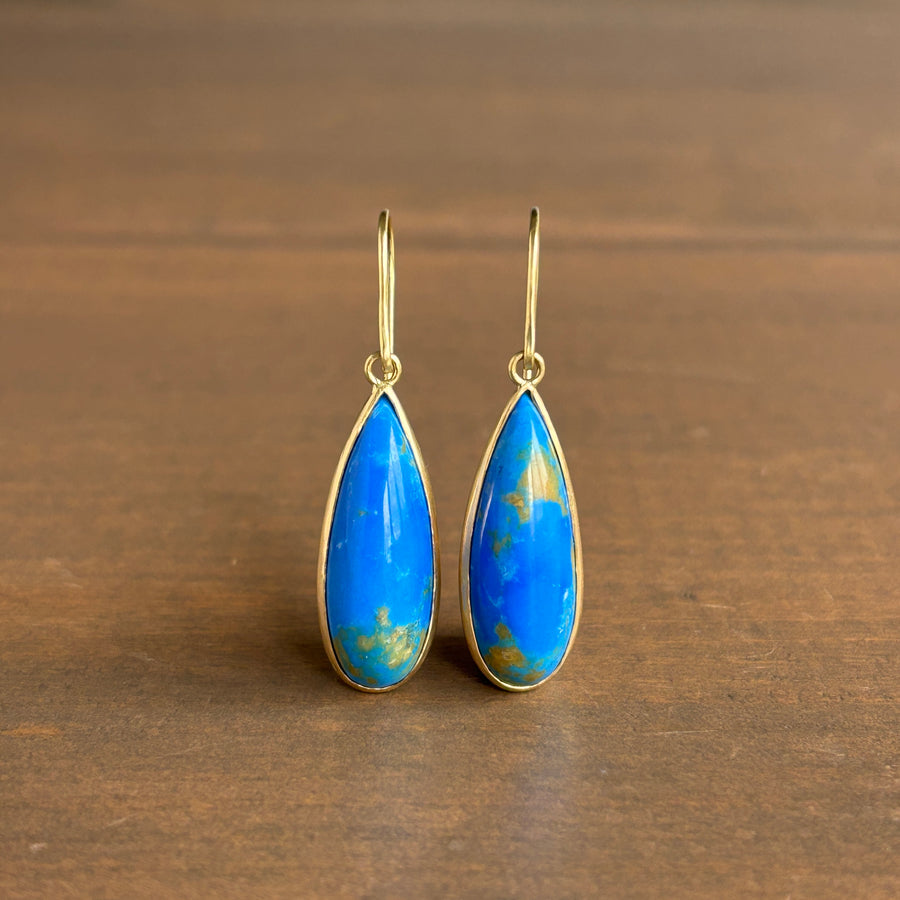 A pair of gold drop earrings with teardrop-shaped blue Ceruleite stones, displayed on a dark brown wooden surface.