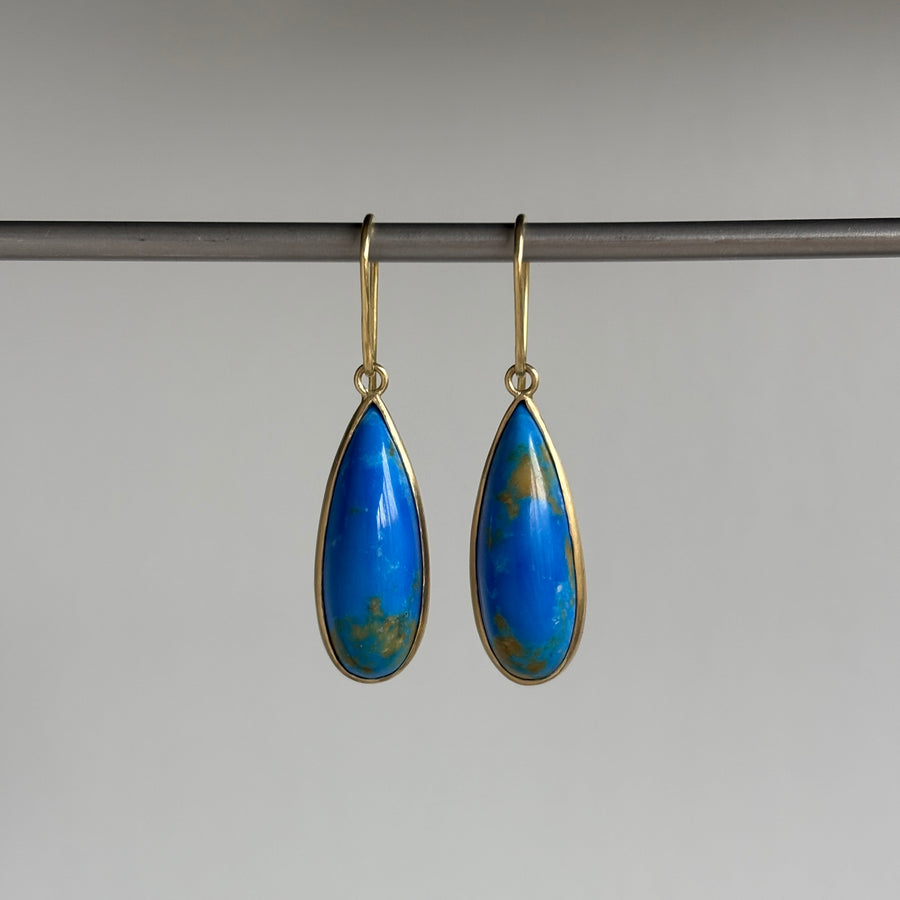 A pair of gold-tone teardrop dangle earrings with vibrant blue Ceruleite inlays, hanging from a dark metal rod against a neutral gray background.