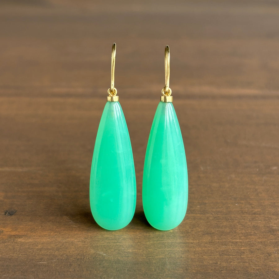 A pair of teardrop-shaped drop earrings made of smooth mint green stone with gold-tone hooks, displayed on a dark brown wooden surface.
