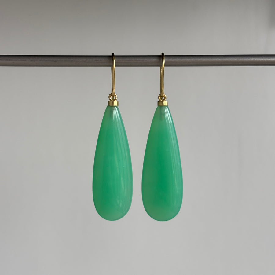A pair of green teardrop-shaped drop earrings with gold fishhook ear wires, hanging from a dark horizontal bar against a plain light gray background.