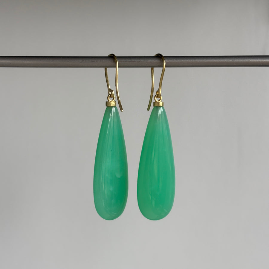 A pair of green teardrop-shaped drop earrings with gold fishhook wires, hanging from a dark horizontal bar against a plain light gray background.