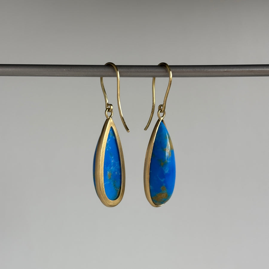 A pair of gold-tone teardrop dangle earrings with vibrant blue Ceruleite inlays, hanging from a dark metal rod against a neutral gray background.