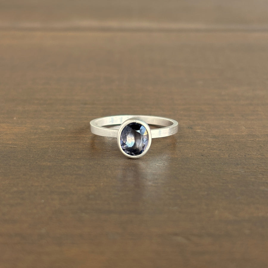 A silver ring with an oval-cut purple gemstone, resting on a brown wooden surface.