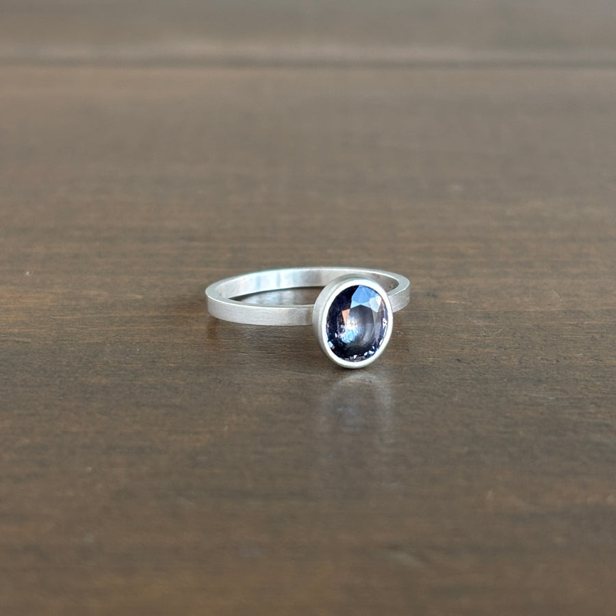 A silver ring with an oval purple gemstone, resting on a dark brown wooden surface.