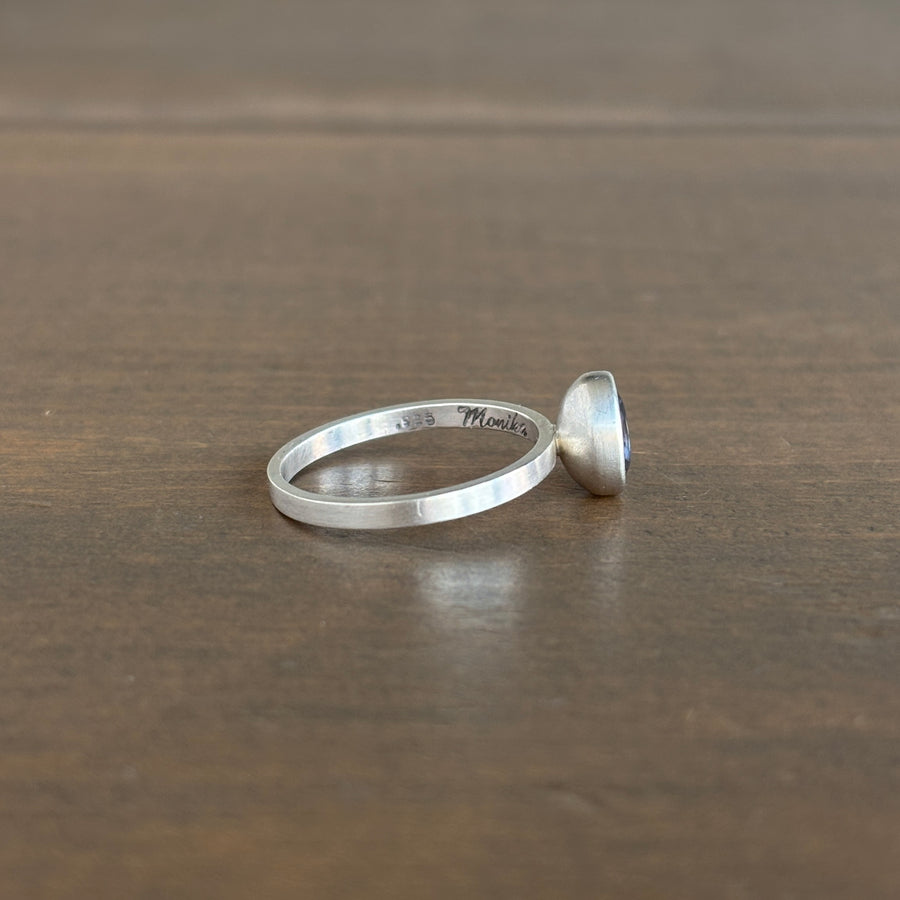 A sterling silver ring with a flat band and a small purple stone, resting on a dark brown wooden surface. 