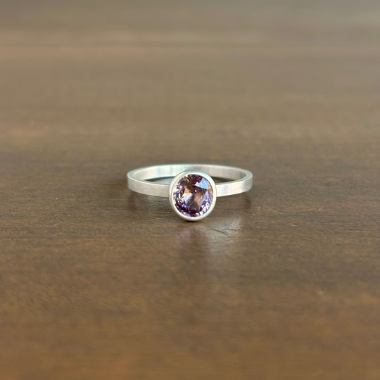 A silver ring with a round bezel-set purple gemstone, resting on a dark brown wooden surface.