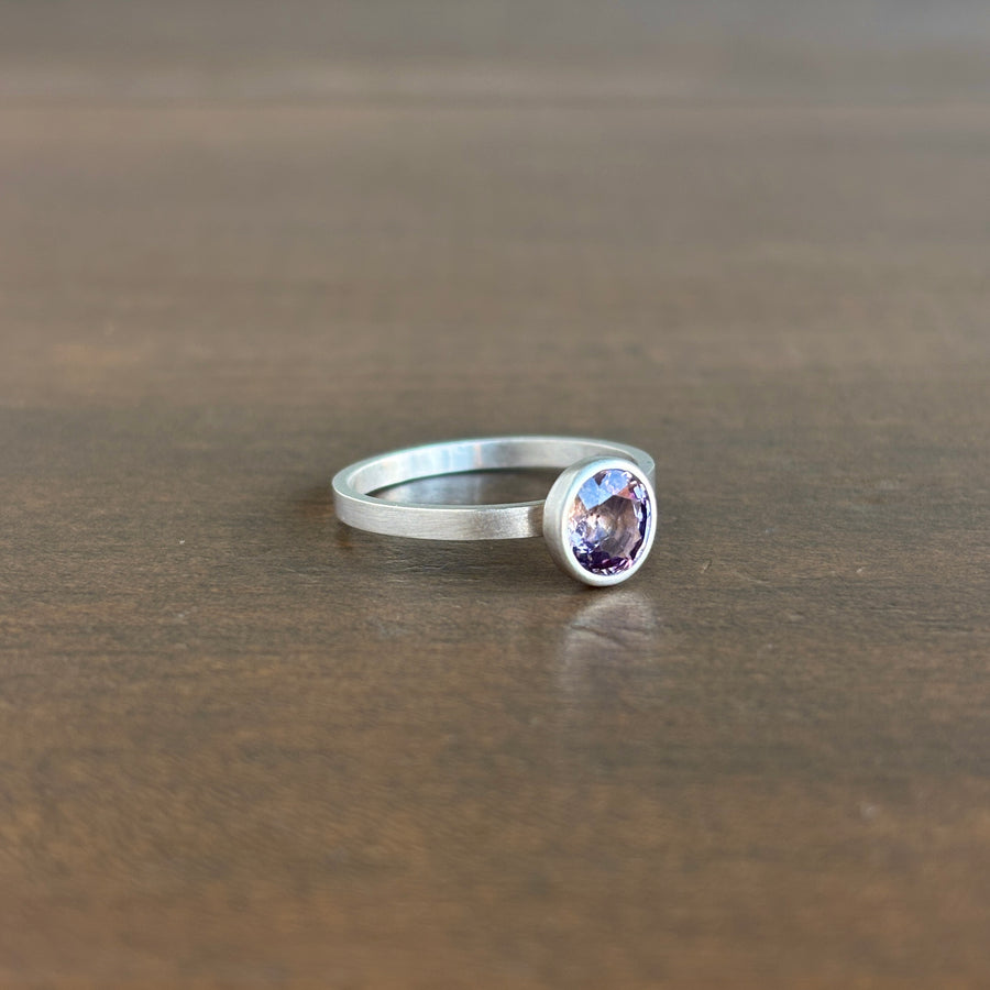 A silver ring with a bezel-set oval purple gemstone, resting on a dark brown wooden surface.
