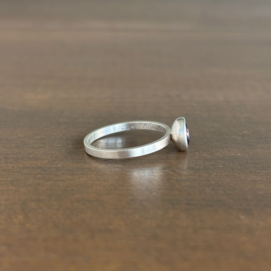A silver band ring with a small, dome-shaped purple gemstone, shown in profile on a dark brown wooden surface.