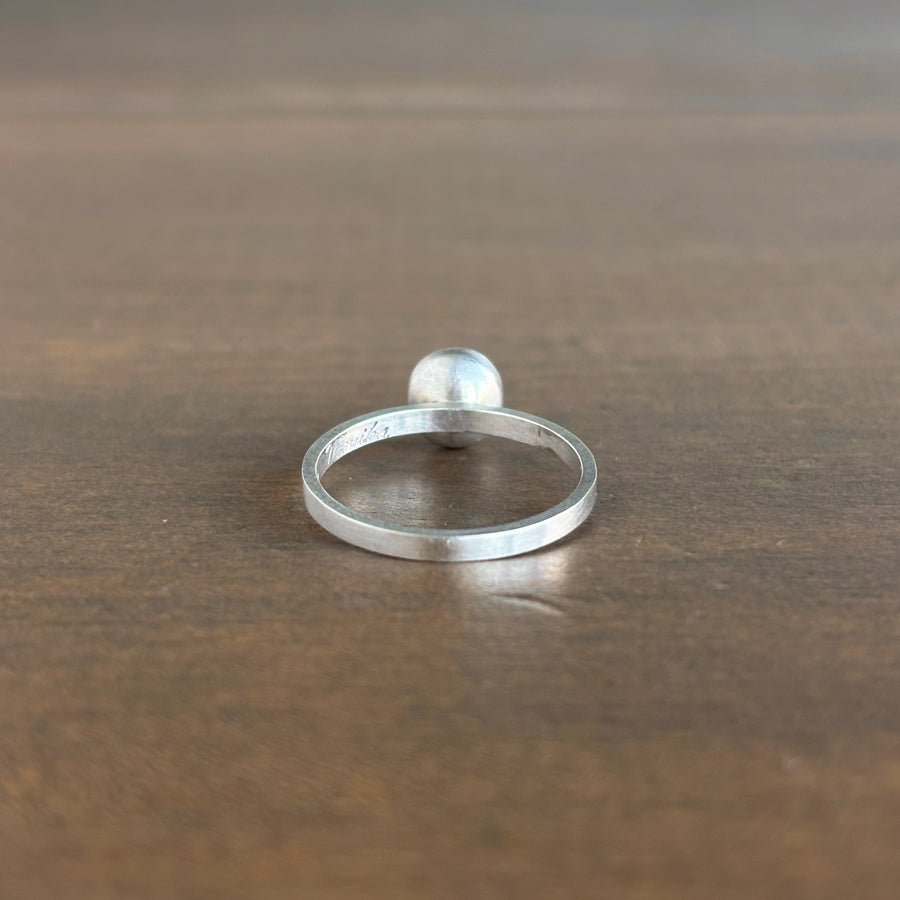 A minimalist sterling silver ring with a flat band and a small, polished round stone, resting on a dark brown wooden surface.