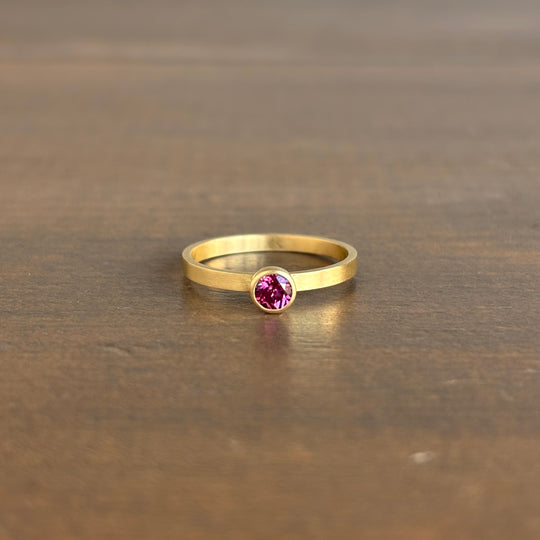 A gold ring with a round pink gemstone set in a bezel setting, resting on a dark brown wooden surface.