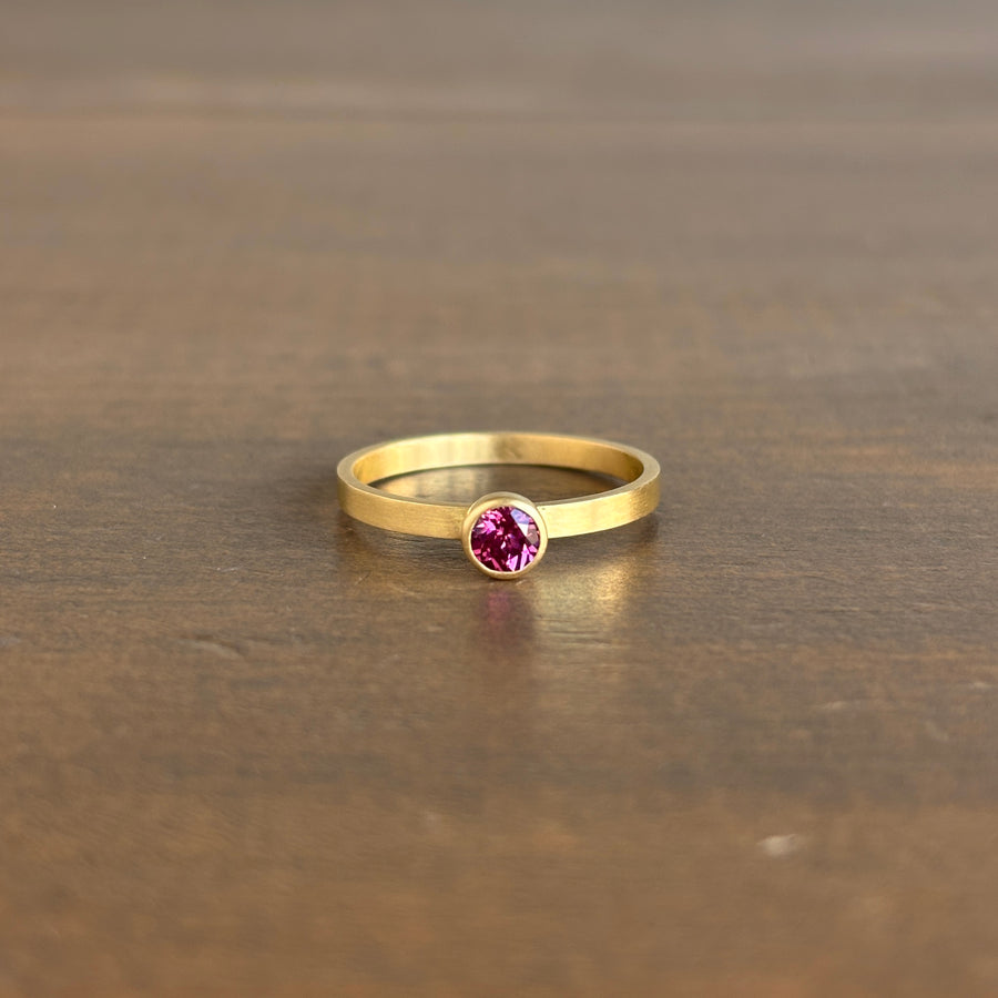 A gold ring with a round pink gemstone set in a bezel setting, resting on a dark brown wooden surface.