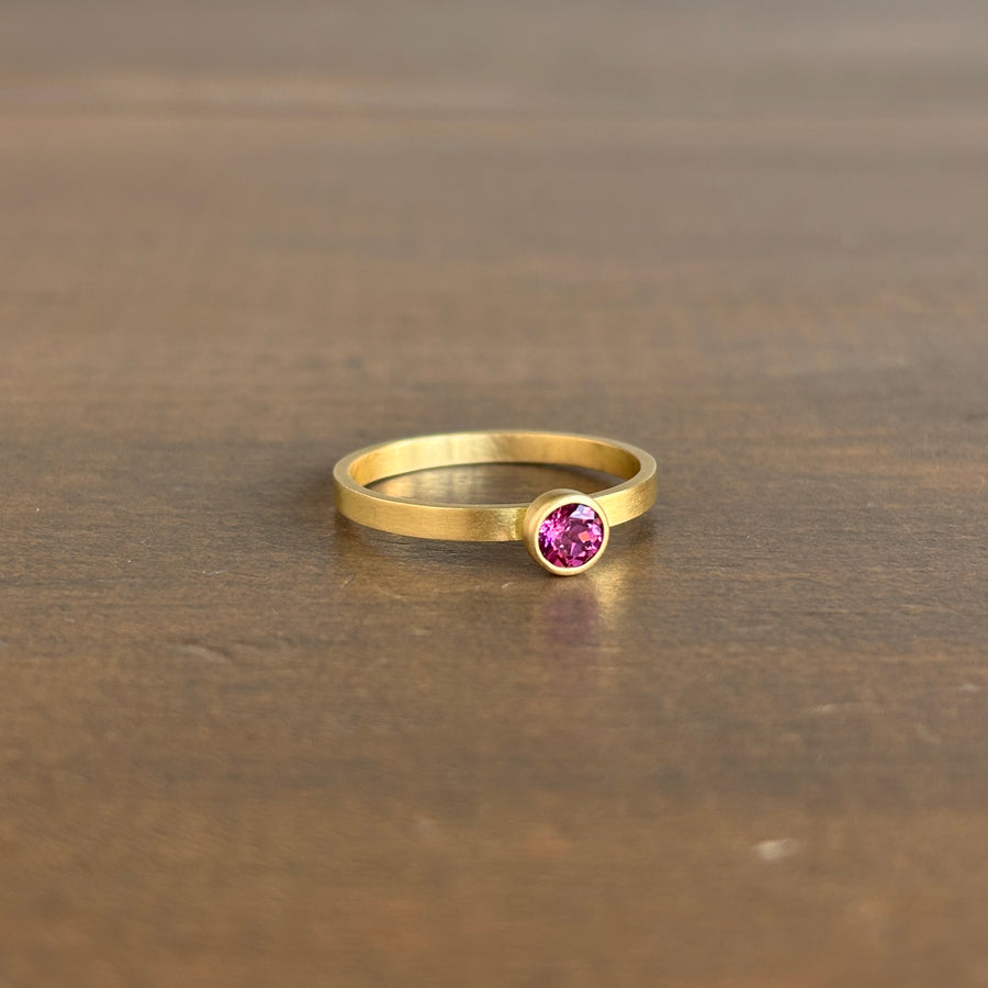 A gold band ring with a single round pink gemstone, resting on a dark brown wooden surface.