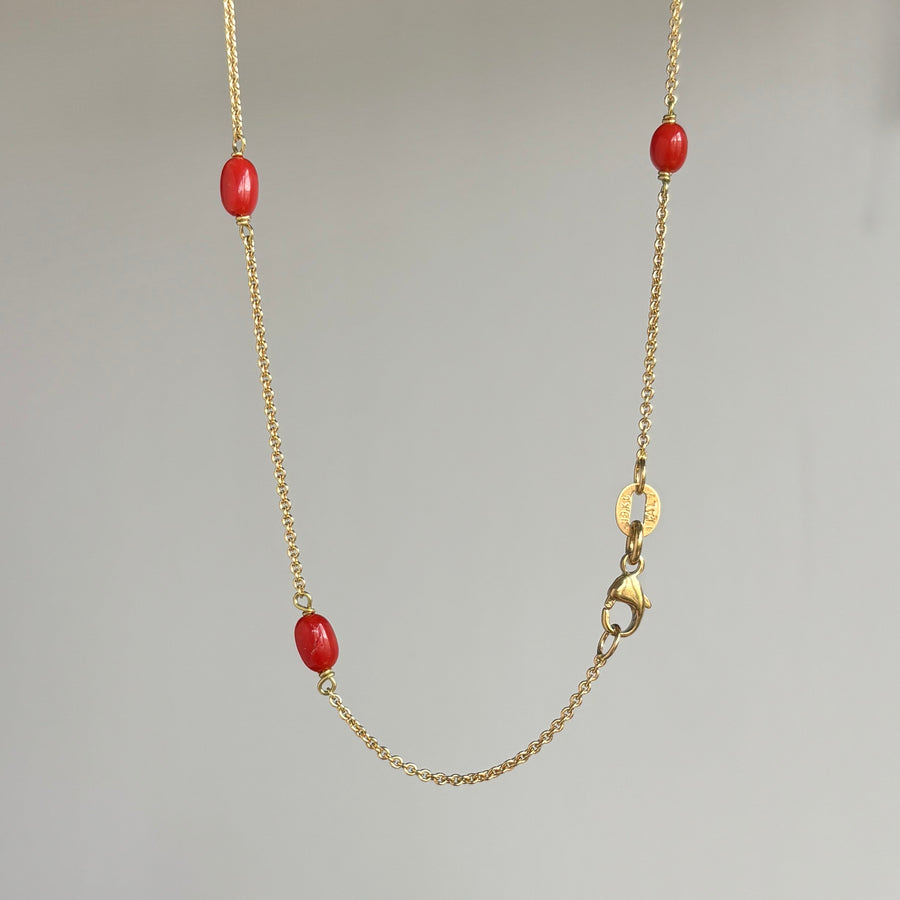 A delicate gold chain necklace featuring three small, polished red oval beads spaced along the chain, displayed against a neutral light gray background.
