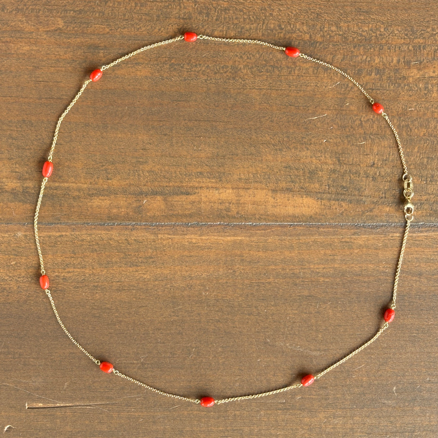 A delicate gold chain necklace with small, polished red coral beads spaced evenly along the chain, laid out in a circle on a rustic brown wooden surface.