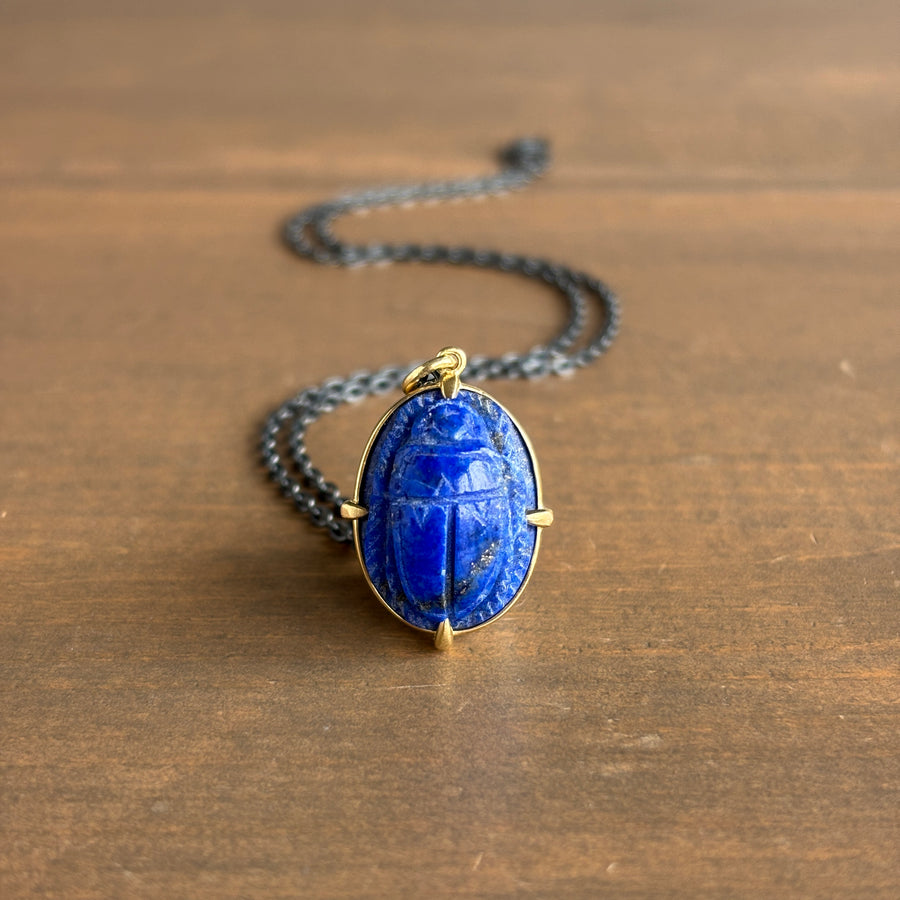 A gold-toned necklace featuring an oval blue lapis lazuli pendant carved with a scarab beetle motif, resting on a brown wooden surface.