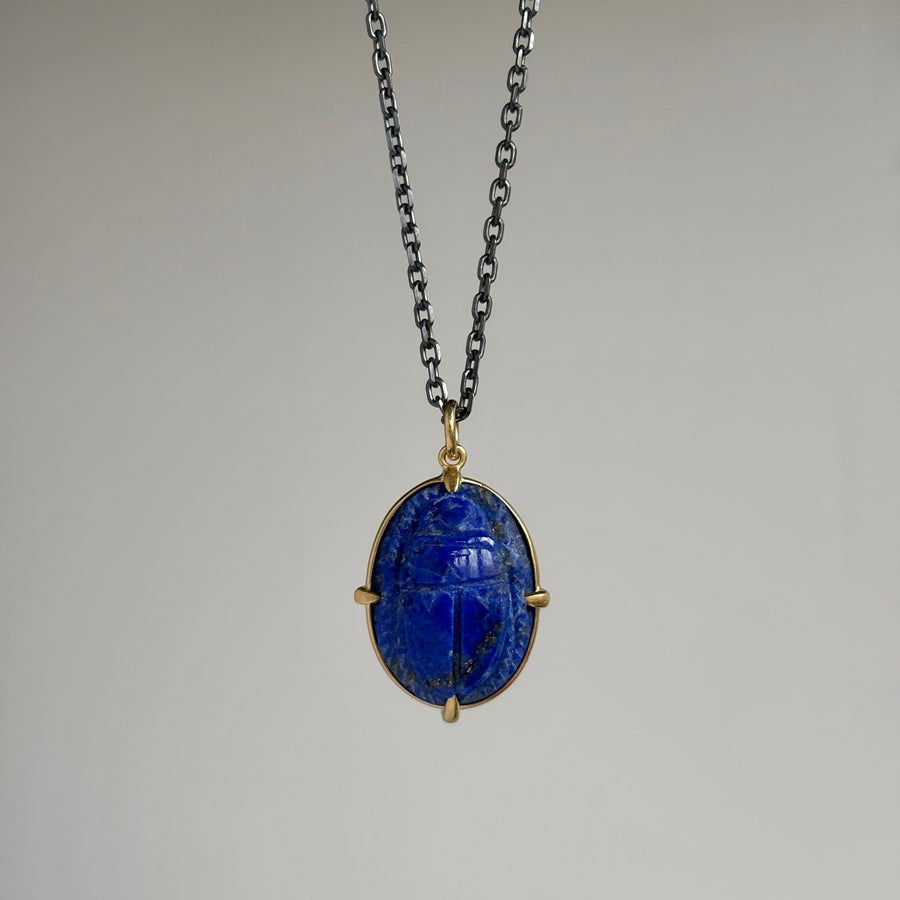 A blue lapis lazuli scarab pendant necklace with gold-toned metal settings and a dark silver chain, shown against a neutral gray background.