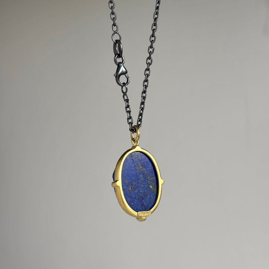 A gold and black chain necklace featuring an oval pendant with a deep blue lapis lazuli stone, shown against a neutral gray background.