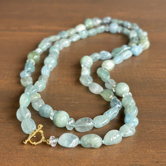 A double-strand beaded necklace made of light blue-green polished stones, featuring a gold toggle clasp and a clear faceted accent bead, displayed on a brown wooden surface.