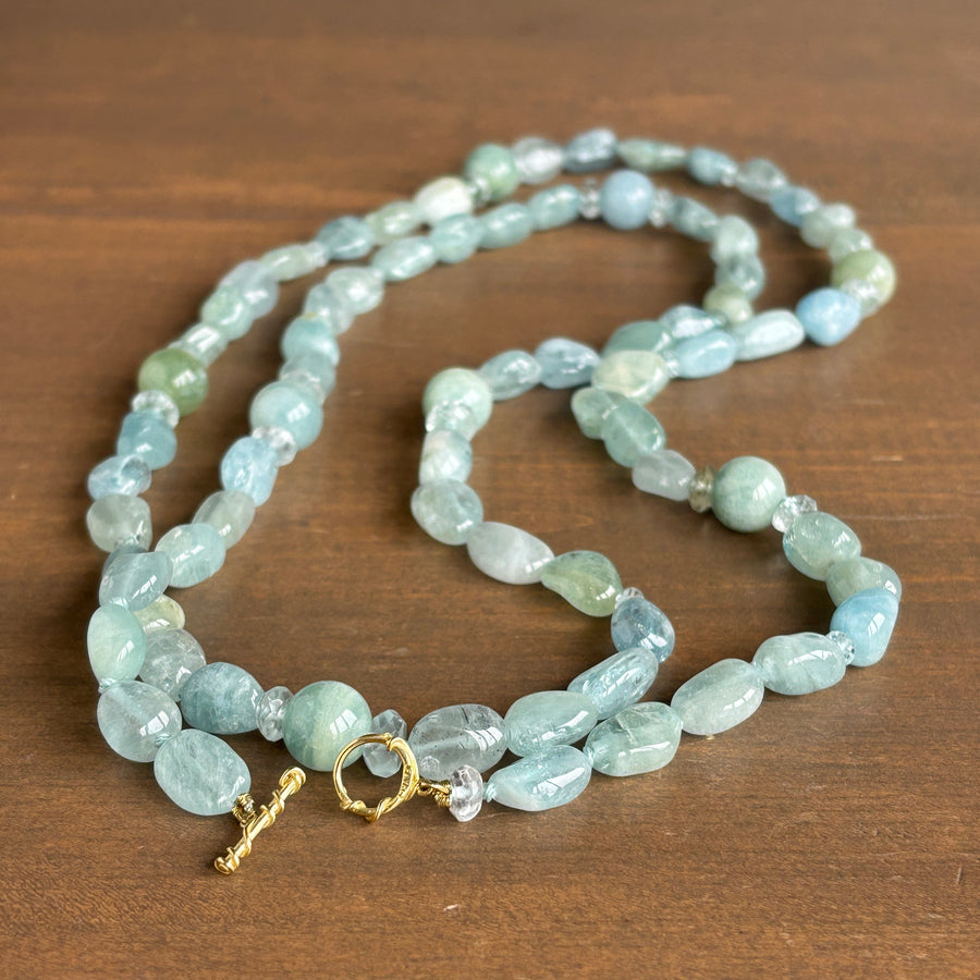 A beaded necklace made of light blue and green stone beads with a small gold clasp, displayed on a brown wooden surface.