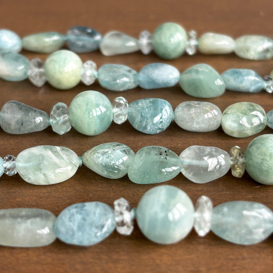 A close-up of a beaded strand featuring light blue and green Aquamarine beads alongside clear faceted crystal spacers, laid out on a brown wooden surface.