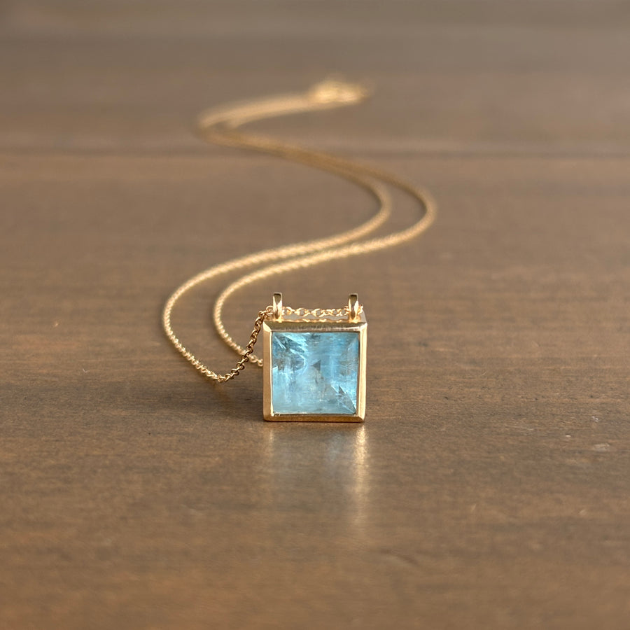 Gold necklace with a square pendant featuring a blue stone on a brown surface