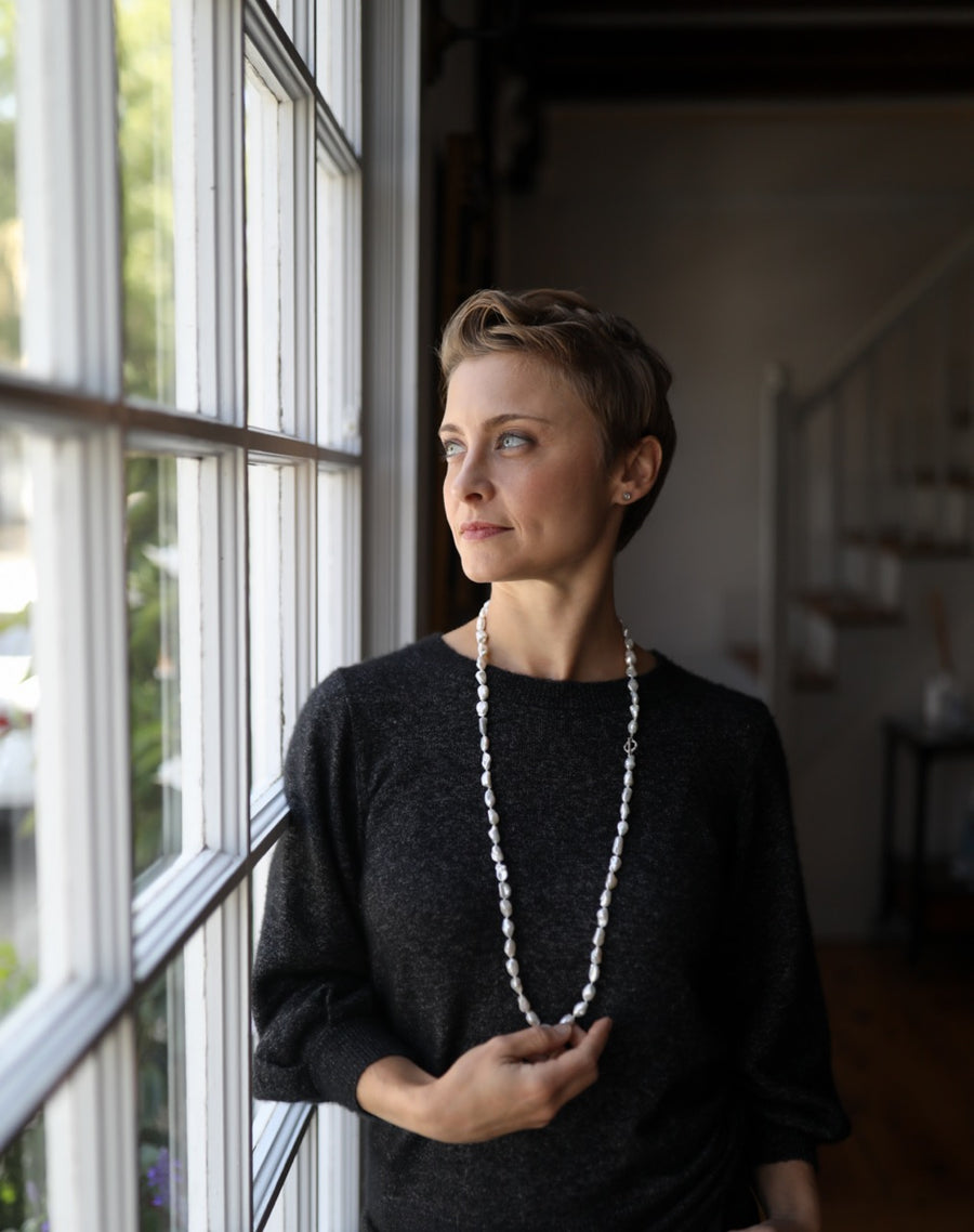 Woman standing by a window holding a long necklace