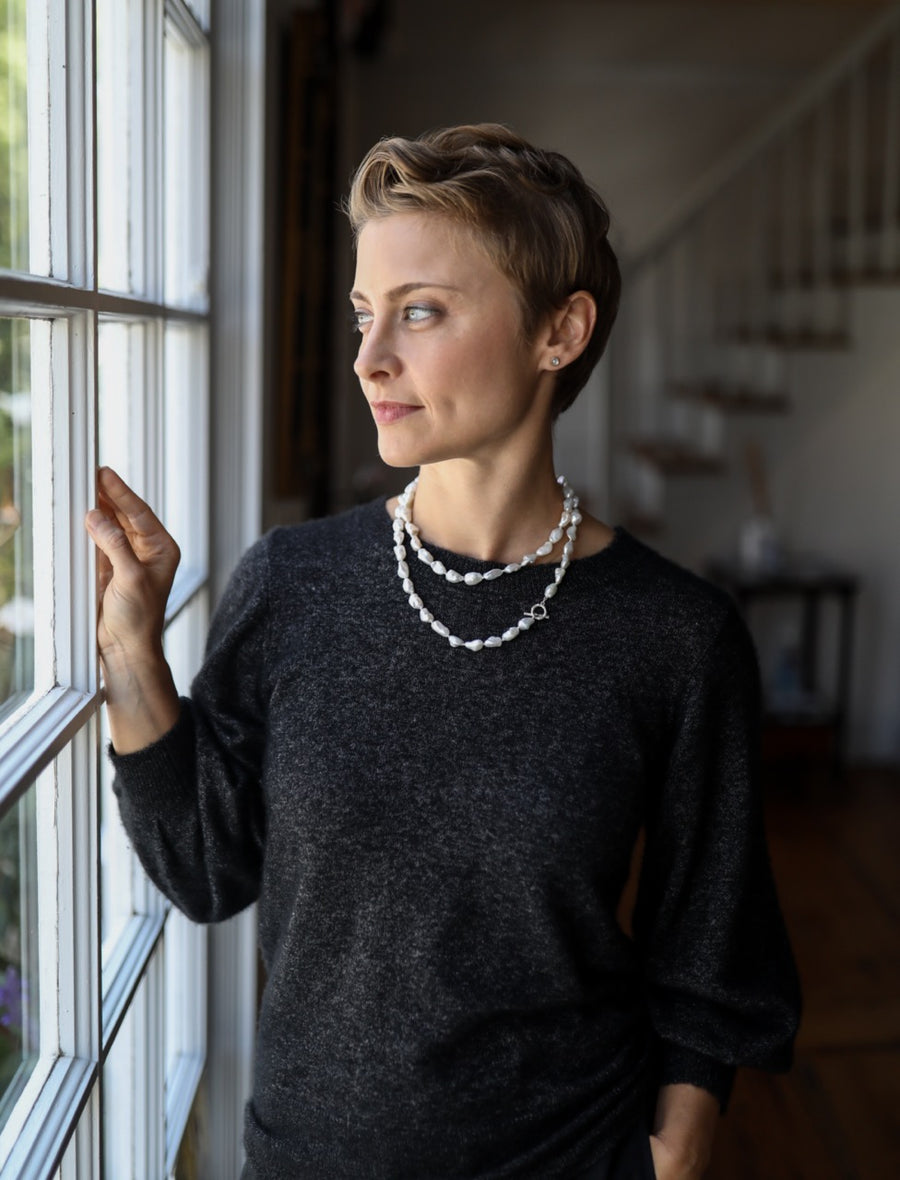Woman standing by a window wearing a dark sweater and pearl necklace.