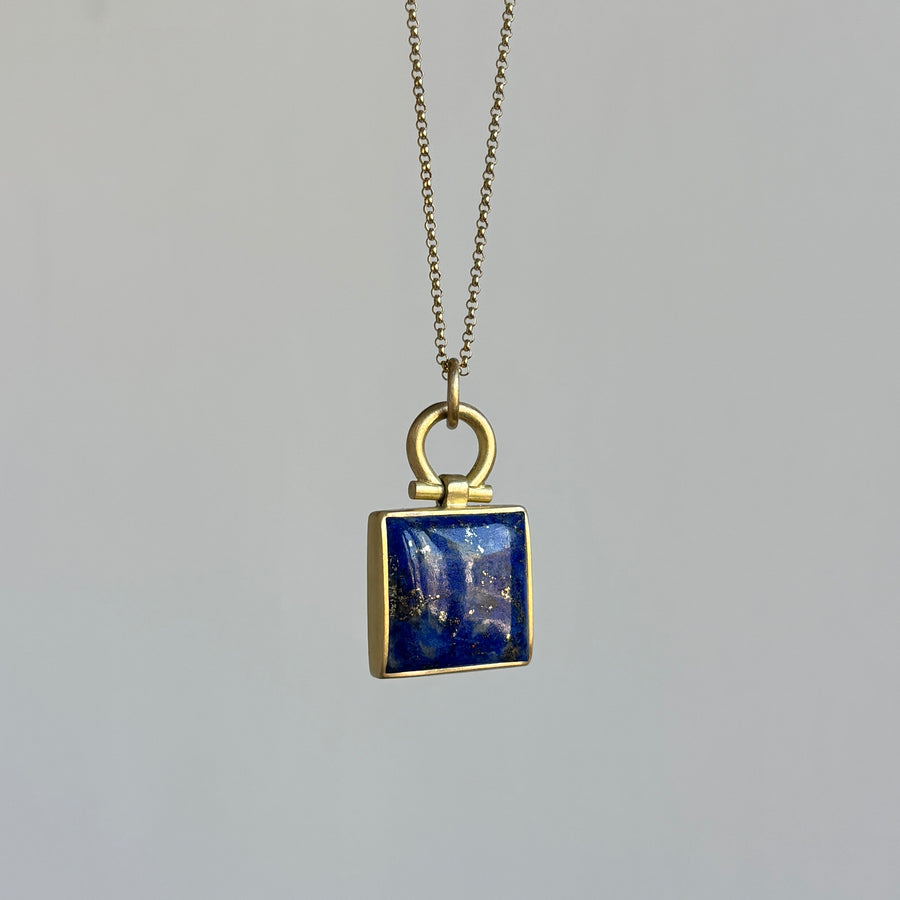 Gold necklace with a square pendant featuring a blue stone on a gray background