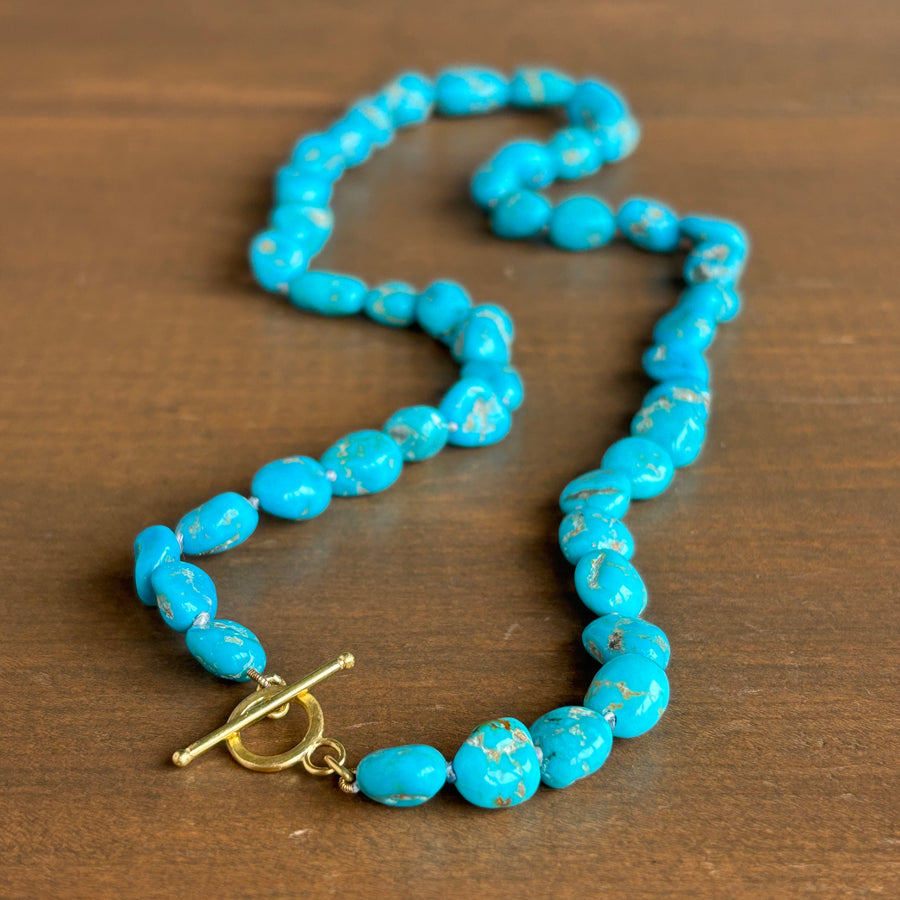 A beaded necklace made of irregular bright turquoise-blue beads with small white marbling, arranged on a brown wooden surface. The necklace has a gold-tone toggle clasp.