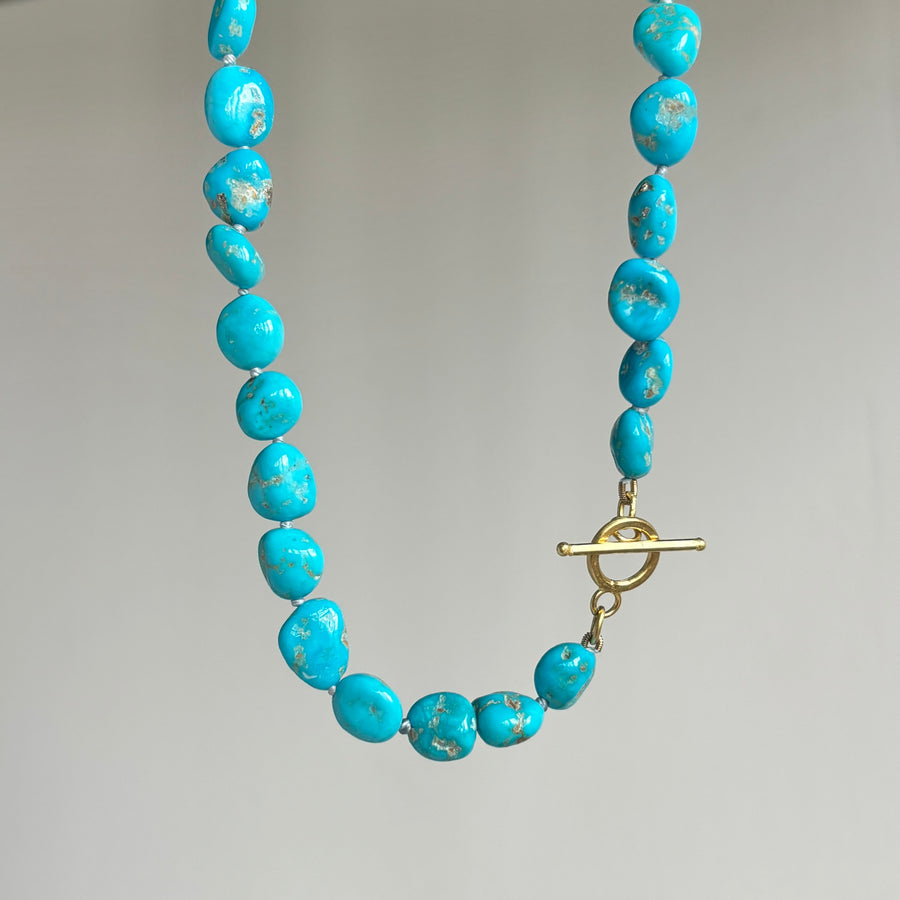 A turquoise bead necklace with a gold toggle clasp, shown against a neutral light gray background.