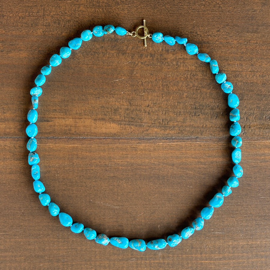 A beaded necklace made of irregular turquoise-blue stones arranged in a circle, with a small gold-tone clasp, photographed on a brown wooden surface.