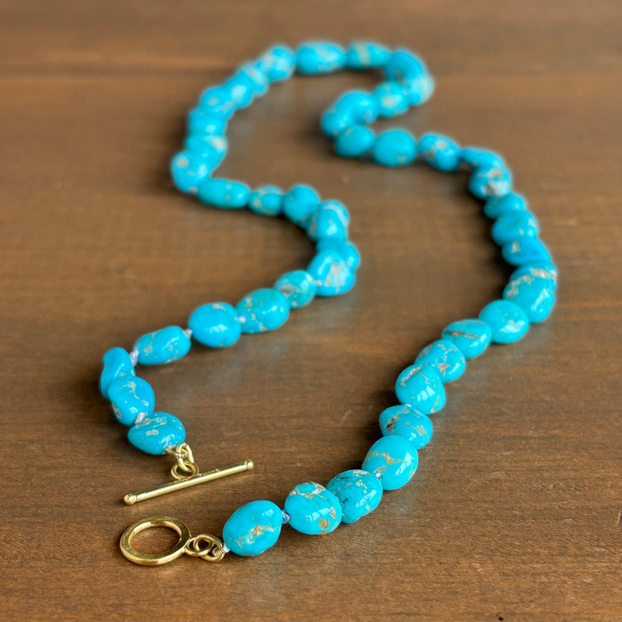 A turquoise beaded necklace with a gold toggle clasp, displayed on a dark brown wooden surface.