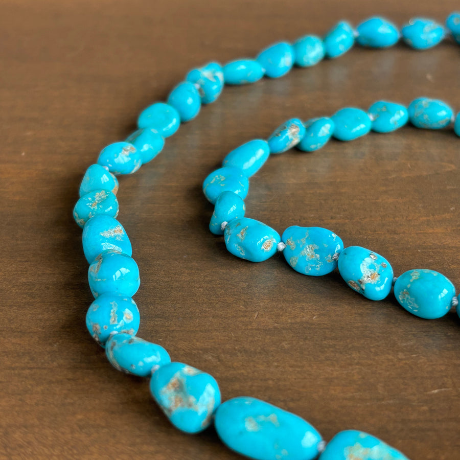 A close-up of a turquoise bead strand or necklace laid on a brown wooden surface. The beads are bright turquoise blue with natural white matrix veining and some small brownish inclusions.
