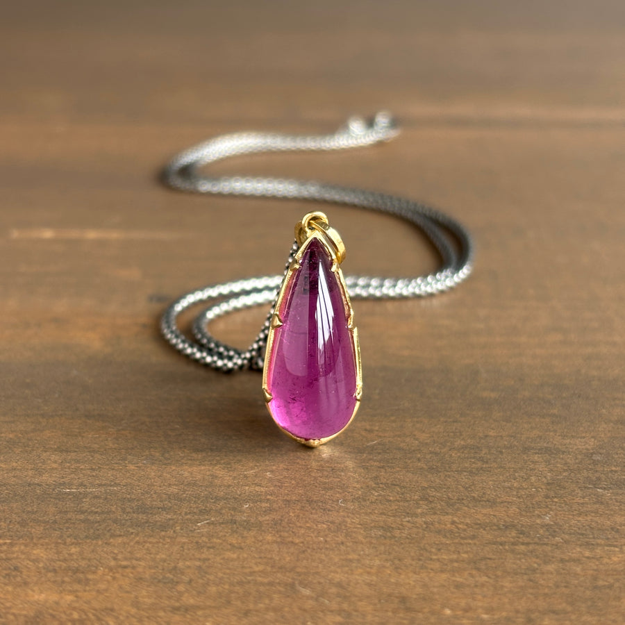 A teardrop-shaped pink gemstone pendant in a gold bezel setting, suspended from a silver chain, resting on a brown wooden surface.