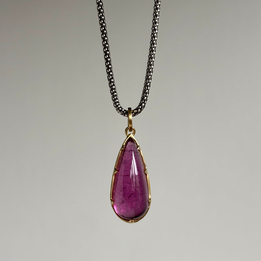 A teardrop-shaped purple gemstone pendant in a gold bezel setting, hanging from a dark silver chain against a neutral gray background.