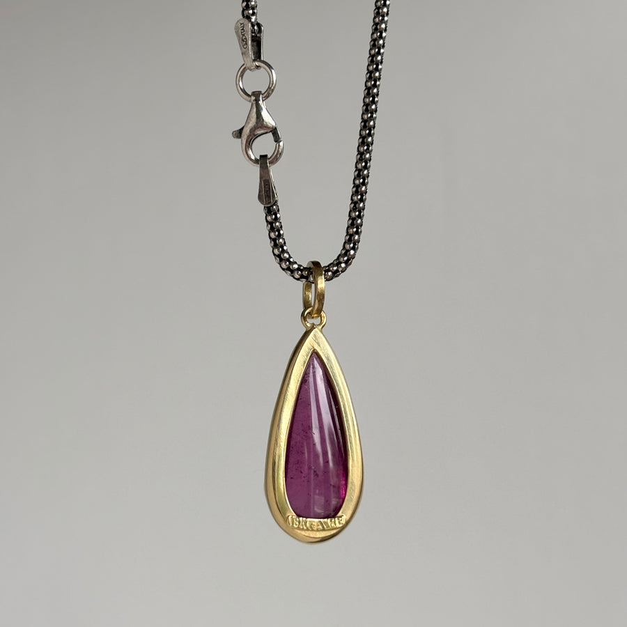 A teardrop-shaped pendant necklace featuring a polished purple gemstone in a gold bezel setting, suspended from a black chain with a silver lobster clasp, shown against a neutral gray background.