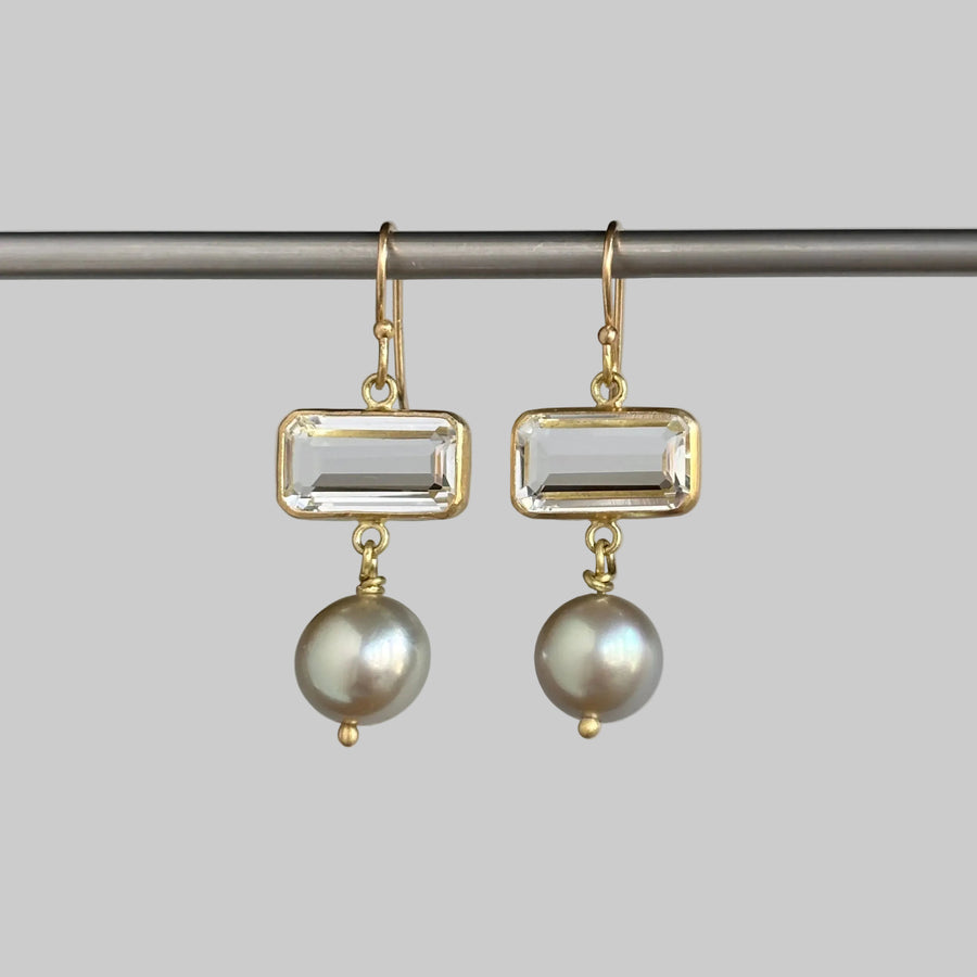 A pair of gold drop earrings featuring a rectangular clear gemstone and a dangling baroque pearl, hanging from a dark horizontal bar against a solid gray background.
