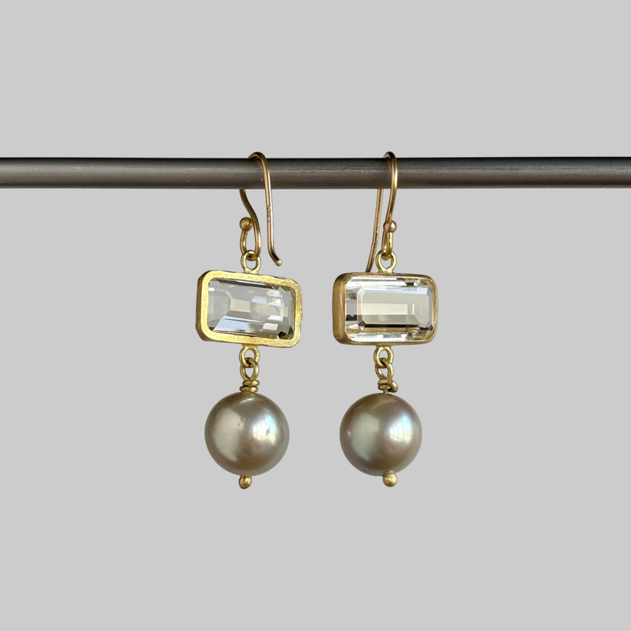 A pair of gold drop earrings featuring rectangular clear crystals and round gray pearls, hanging from a dark horizontal bar against a solid gray background.