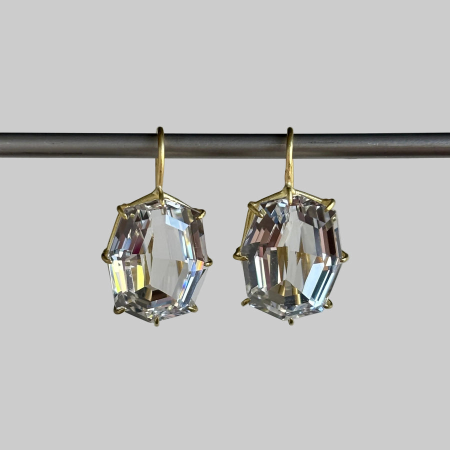 A pair of gold-tone drop earrings with large, faceted clear crystal gemstones, hanging from a dark horizontal bar against a light gray background.