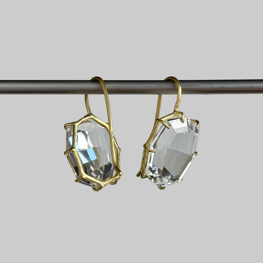 A pair of gold-toned drop earrings with large, faceted clear gemstones, hanging from a dark metal rod against a solid gray background.