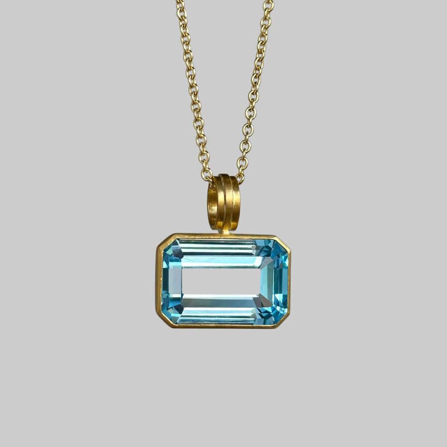 A gold chain necklace with a rectangular light blue gemstone pendant in a gold bezel setting, shown against a neutral light gray background.